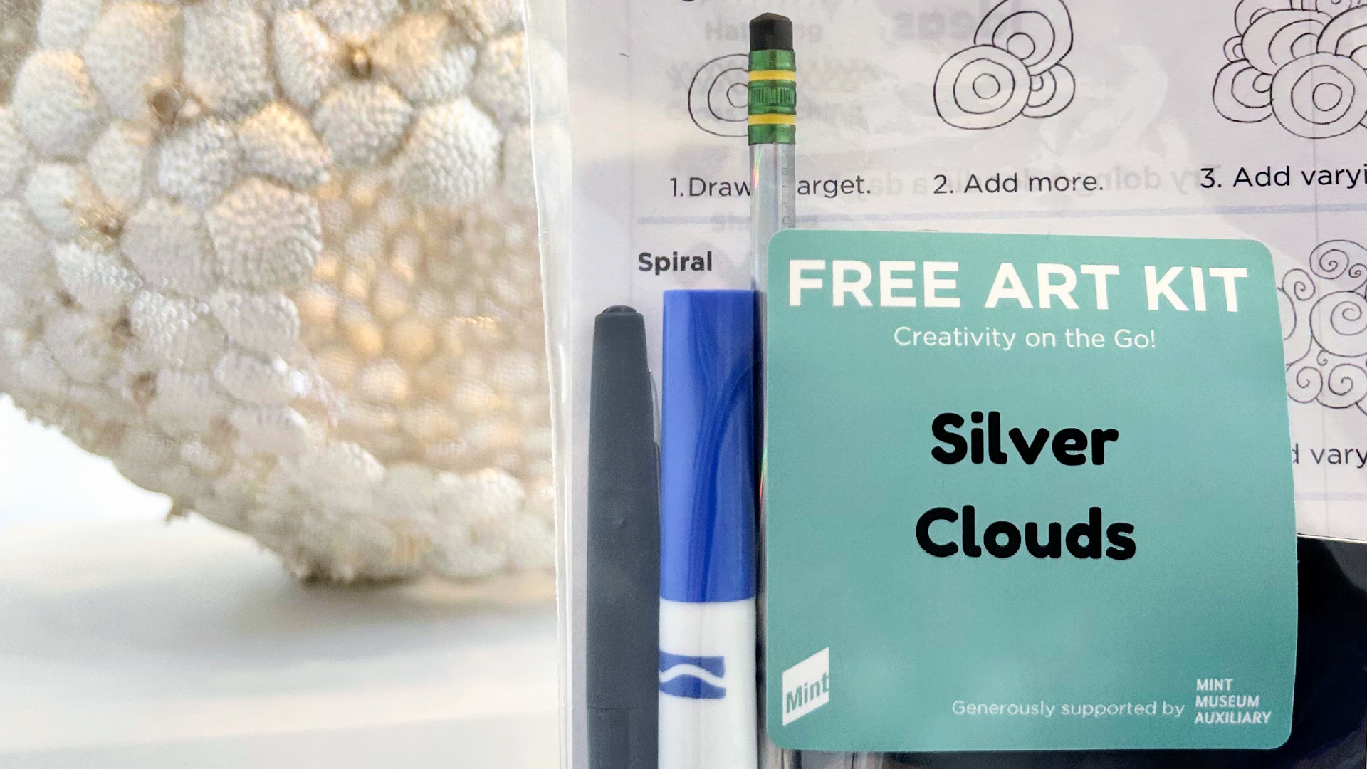 Photo of a free art kit being held in front of a cloud-like sculpture made of silver.