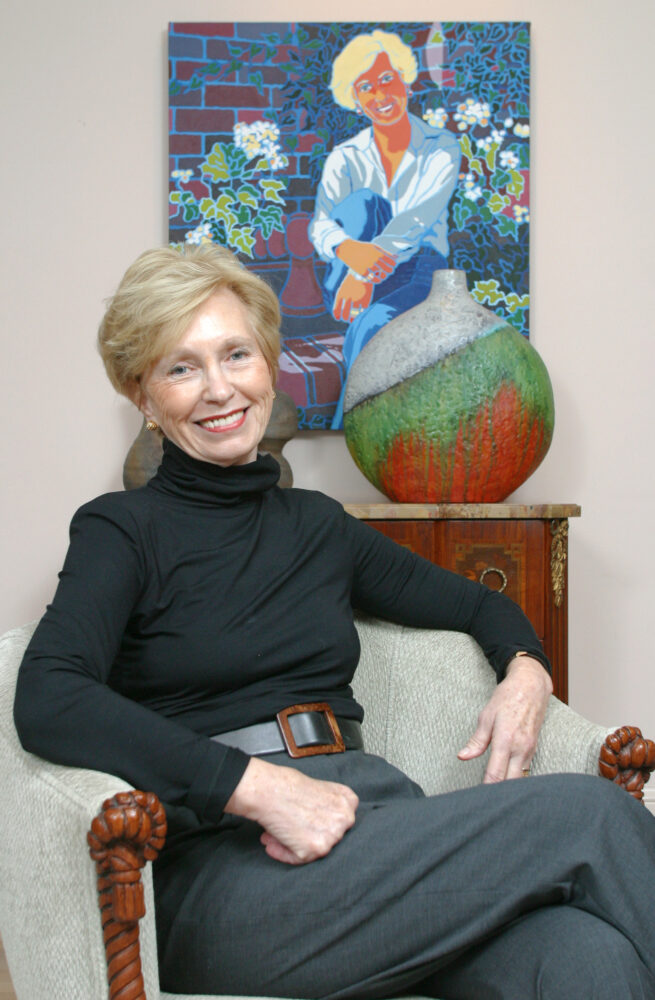 Woman with fair skin and blonde hair dressed in a black turtleneck and gray pants sitting in front of a portrait.